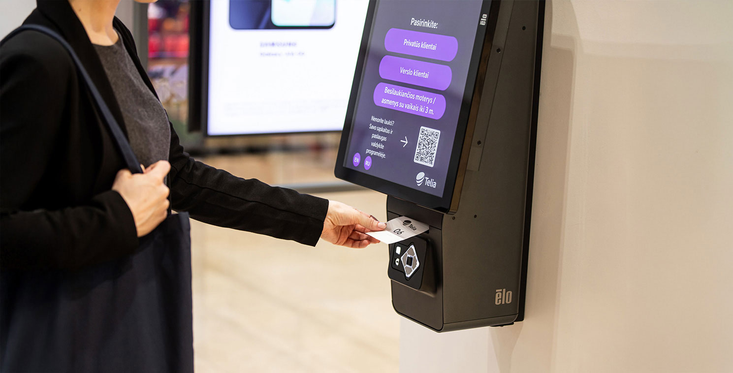 A Telia customer retrieves a printed queue ticket from an Elo touchscreen self service kiosk, streamlining in-store check in and service management