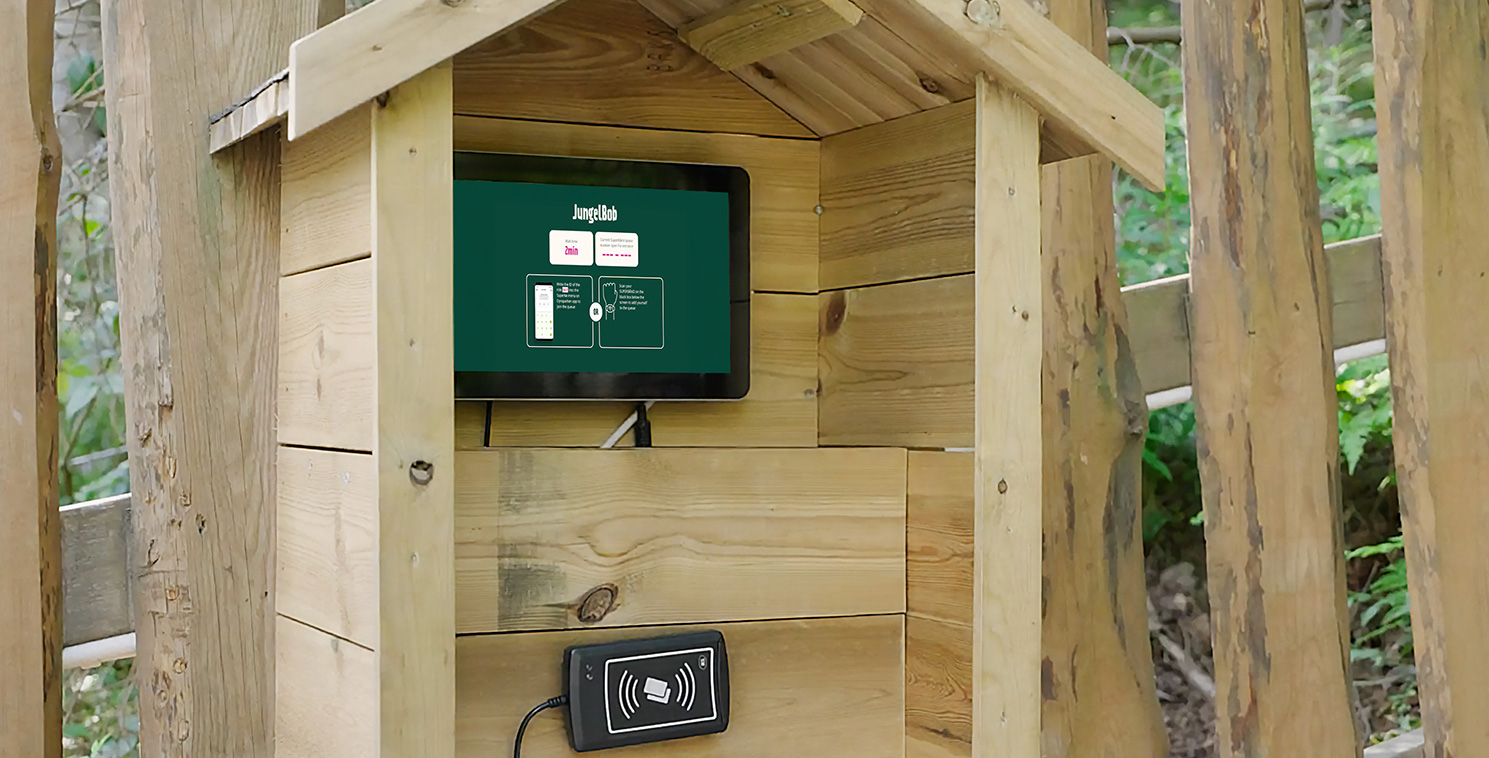 Un visiteur utilise une borne tactile résistante aux intempéries installée dans un boîtier en bois dans un parc d’attractions, illustrant la technologie libre-service pour l’achat de billets.